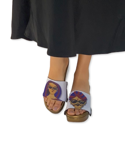 Mid-length shot of slim model wearing Desi Cool ethnic strap sandal with Rajasthani folk art characters in sunglasses and cork wedge, styled with indigo linen trousers in a Jaipur blue city lane, by Soule Footwear